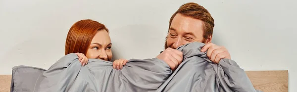 Day off without kids, redhead husband and wife looking at each other, hiding behind blanket, married couple, modern lifestyle, happiness at home, relaxation time, parents alone at home, banner — Stock Photo