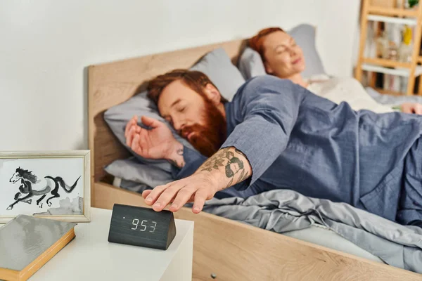 Сонный бородатый мужчина достигает электронный будильник, выходной день без детей, рыжий муж и жена, время релаксации, размытый фон, татуировка, современный дом, уютная спальня — стоковое фото