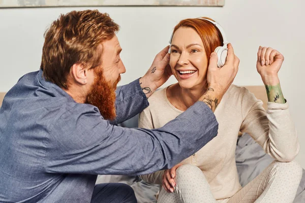 Music enjoyment, quality time, day off without kids, redhead husband and wife, bearded man wearing wireless headphones on woman, cheerful parents alone at home, modern lifestyle, relationship — Stock Photo