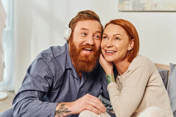 Music lovers, day off without kids, redhead husband and wife, woman leaning on bearded man in wireless headphones, cheerful parents alone at home, modern lifestyle, listening music — Stock Photo