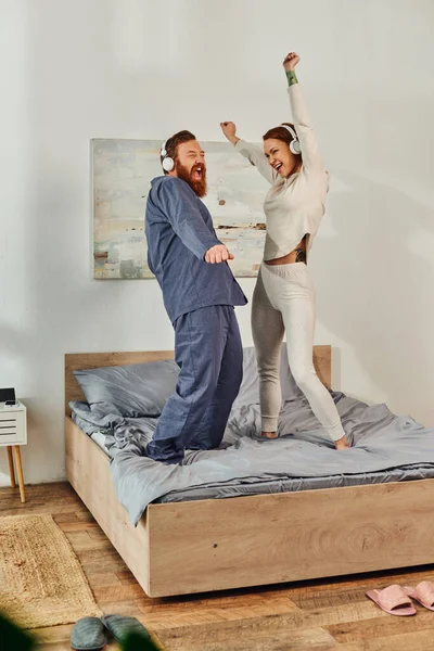 Day off without kids, having fun, shared music, tattooed husband and wife, happy couple in wireless headphones dancing on bed, bearded man and carefree woman, redhead music lovers, weekends — Stock Photo