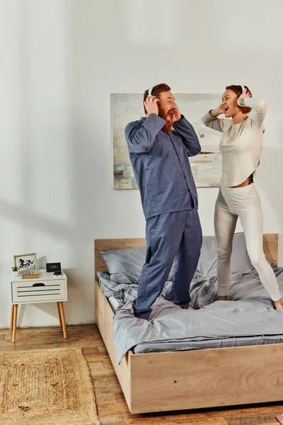 Day off without kids, having fun, shared music, excited husband and wife, happy couple in wireless headphones dancing on bed, bearded man and redhead woman, tattooed music lovers, weekends — Stock Photo