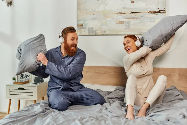 Day off without kids, having fun, shared music, redhead husband and wife, happy couple in wireless headphones pillow fight, bearded man and carefree woman, tattooed music lovers, weekends — Stock Photo