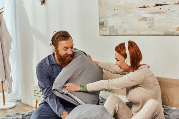 Parents alone, having fun, shared music, redhead husband and wife, happy couple in wireless headphones pillow fight, bearded man and carefree woman, tattooed people, day off without kids — Stock Photo