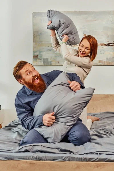 Parents alone, having fun, pillow fight, redhead husband and wife, happy couple in sleepwear enjoying time together, bearded man and carefree woman, tattooed people, day off without kids — Stock Photo