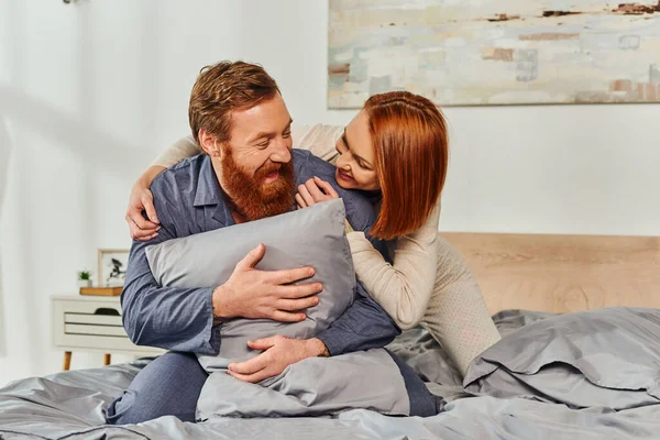 Выходной день без детей, рыжая женщина обнимает бородатого мужа подушкой, счастливая пара во сне наслаждается временем вместе, татуированные люди, родители одни дома, качественное время вместе — стоковое фото