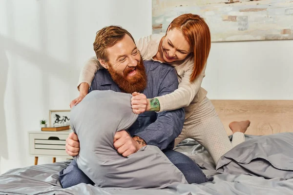 Выходной день без детей, рыжая женщина обнимает бородатого мужа подушкой, счастливая пара во сне наслаждается временем вместе, татуированные люди, родители одни дома, качественное время вместе, игривый — стоковое фото