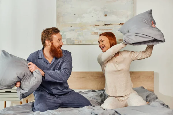 Day off without kids, having fun, pillow fight, redhead husband and wife, happy couple in sleepwear enjoying time together, bearded man and carefree woman, tattooed people — Stock Photo
