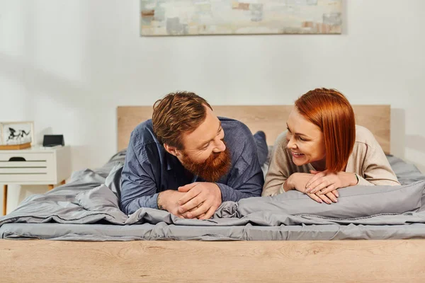 Day off without kids, happy couple relaxing together, lying on bed, redhead husband and wife, enjoying time together,  bearded man and carefree woman looking at each other, tattooed people — Stock Photo