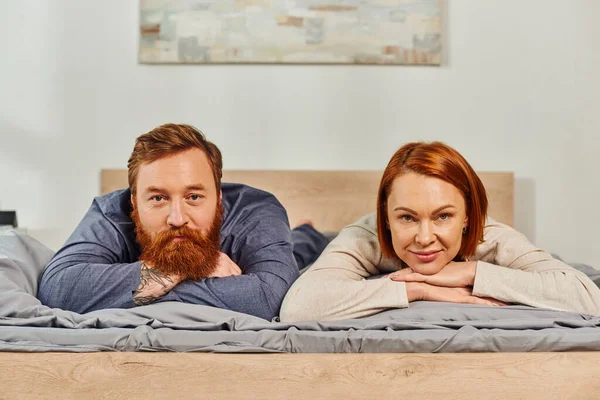 Jour de congé sans enfants, couple heureux se détendre ensemble, couché sur le lit, rousse mari et femme, profiter du temps ensemble, homme barbu et femme insouciante regardant la caméra, personnes tatouées — Photo de stock