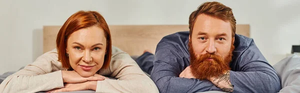 Day off without kids, happy couple relaxing together, lying on bed, redhead husband and wife, enjoying time together,  bearded man and carefree woman looking at camera, tattooed people, banner — Stock Photo