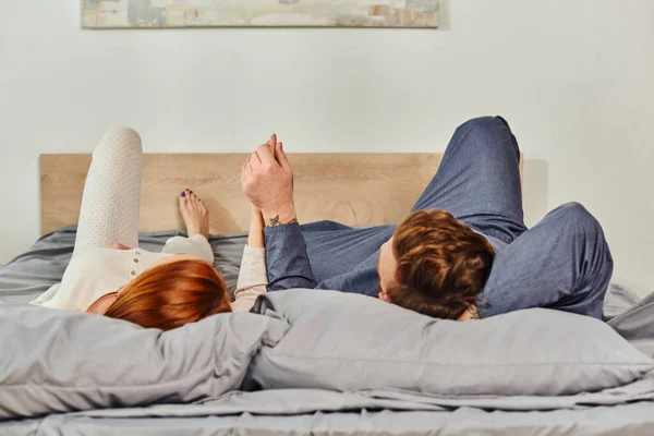 Day off without kids, couple relaxing together, lying on bed, redhead husband and wife, enjoying time together,  bearded man and carefree woman holding hands, tattooed people — Stock Photo