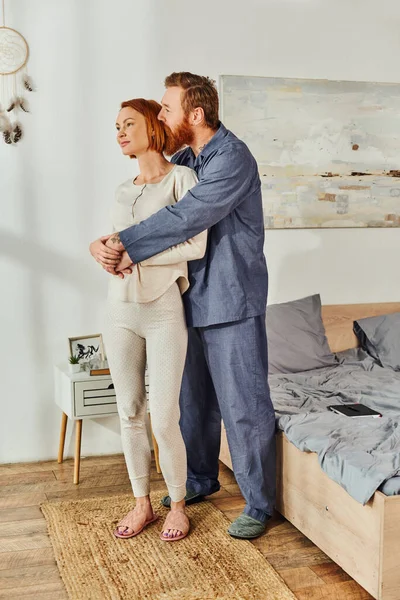 Full length of married couple hugging in cozy bedroom, day off without kids, redhead husband and wife, enjoying time together, weekends together, tattooed, parents alone at home — Stock Photo