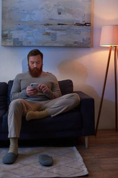 Просмотр телефона, мобильное взаимодействие, бородатый мужчина с рыжими волосами с помощью смартфона, сидя на диване живопись на стене, тапочки на ковре, ночь, свет от лампы, досуг, цифровой век — стоковое фото