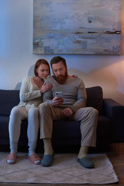 Social media, night, redhead couple spending time without kids, day off, husband and wife, bearded man using smartphone near woman, comfortable living, cozy living room, carefree, tattooed — Stock Photo