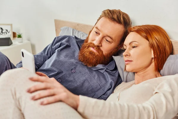 Weekends without kids, morning, relaxation time, tattooed couple relaxing in cozy bedroom, husband and wife, bearded man and redhead woman using smartphone, carefree, screen time, day off — Stock Photo