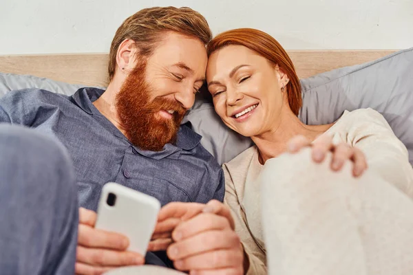 Weekends without kids, morning, relaxation time, tattooed couple relaxing in cozy bedroom, husband and wife, bearded man using smartphone near redhead woman, carefree, screen time, day off — Stock Photo