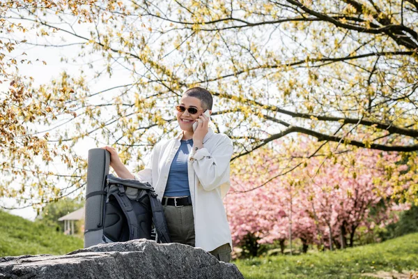 Улыбающаяся коротковолосая и татуированная туристка в солнечных очках, разговаривающая по смартфону и стоящая рядом с рюкзаком на камне с живописной природой на заднем плане, уверенная женщина-исследователь — стоковое фото