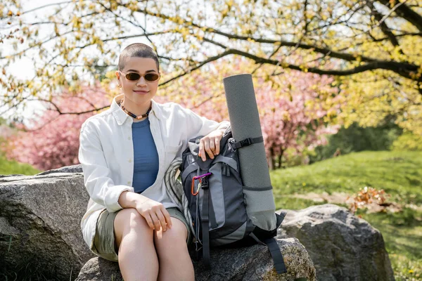 Положительный короткие волосы и молодая женщина турист в повседневной одежде и солнцезащитные очки сидя рядом рюкзак с фитнес-мат на камне с размытой природой на заднем плане, уверенный женщина исследователь — стоковое фото