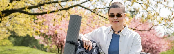 Positive young and short haired female backpacker in casual clothes and sunglasses standing near backpack with fitness mat with nature at background, confident female explorer, banner — Stock Photo