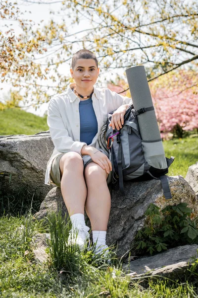 Young short haired and tattooed female traveler looking at camera while sitting near backpack and fitness mat on stone with blurred landscape at background, Translation of tattoo: love — Stock Photo
