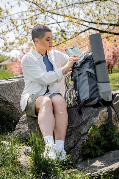 Молода короткошерста і татуйована жінка, що ходить в повсякденному одязі, беручи карту з рюкзака з фітнес-мате, сидячи на камені з розмитою природою на фоні, впевнена жінка-дослідник — стокове фото