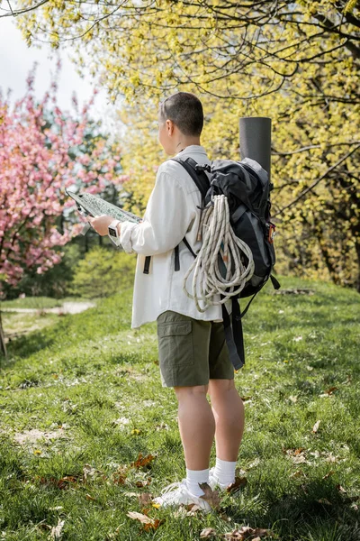Young short haired and tattooed female tourist with backpack, fitness mat and travel equipment while holding map and standing with nature at background, independent traveler embarking on solo journey — Stock Photo