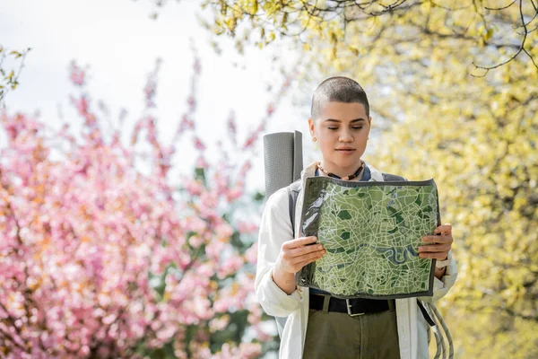 Young short haired female tourist in casual clothes with backpack looking at map while standing with blurred landscape at background, independent traveler embarking on solo journey — Stock Photo