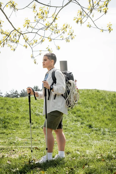 Junge kurzhaarige Reiseleiterin in Freizeitkleidung mit Rucksack und Reiseausrüstung, die mit Trekkingstöcken mit Landschaft im Hintergrund wandert, unabhängige Reisende, die auf Alleinreise geht — Stockfoto