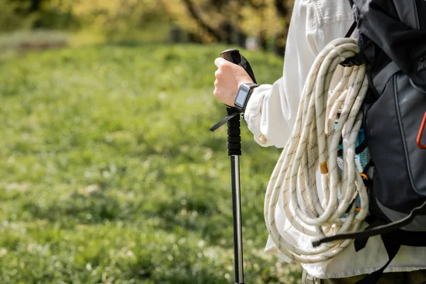 Ausgeschnittene Ansicht einer jungen Wanderin in lässiger Kleidung mit Rucksack, der eine Trekkingstange hält, während sie auf verschwommenem Gras spaziert, unabhängige Reisende, die sich auf eine Alleinreise begeben — Stockfoto