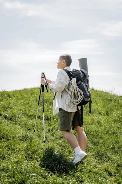 Seitenansicht einer jungen kurzhaarigen Reisenden mit Rucksack und Kletterseil beim Wandern mit Trekkingstöcken auf einem Hügel mit Gras im Hintergrund, unabhängige Reisende auf Alleinreise — Stockfoto