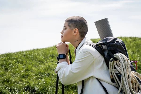Side view of young short haired and tattooed young tourist with backpack holding trekking poles and looking away while standing with hill at background, explorer woman discovering hidden trails — Stock Photo