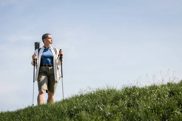 Young short haired woman hiker in casual clothes with backpack holding trekking poles and standing on grassy hill and sky at background, explorer woman discovering hidden trails — Stock Photo