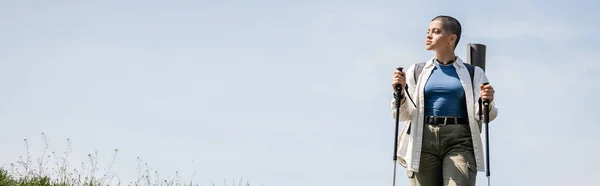 Молодая коротковолосая женщина-путешественница в повседневной одежде с рюкзаком, держа в руках треккинговые шесты, стоя с небом на заднем плане, исследователь женщина обнаруживает скрытые тропы, баннер — стоковое фото