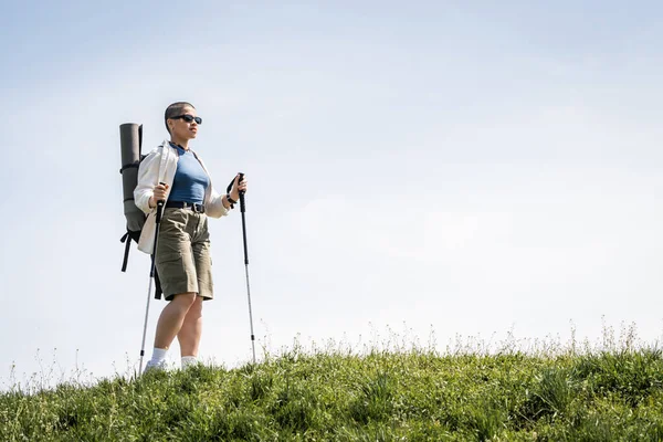 Short haired young female traveler in sunglasses with backpack and travel equipment holding trekking poles while walking on grassy hill , explorer woman discovering hidden trails — Stock Photo