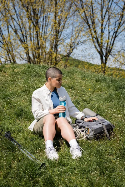Joven viajera femenina de pelo corto sosteniendo botella deportiva y sentada cerca de la mochila y los bastones de trekking en una colina cubierta de hierba con la naturaleza al fondo, exploradora, Traducción del tatuaje: amor - foto de stock