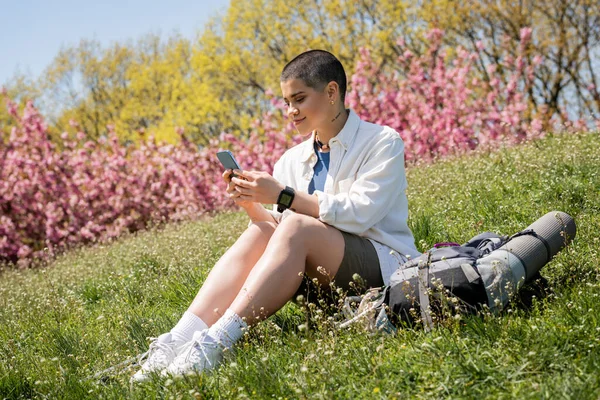 Sonriente joven viajera de pelo corto utilizando un teléfono inteligente mientras está sentada cerca de la mochila con equipo de viaje en una colina cubierta de hierba con la naturaleza en el fondo, excursionista curioso explorando nuevos paisajes - foto de stock
