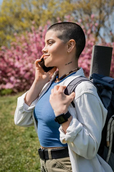 Seitenansicht einer jungen, kurzhaarigen und tätowierten Wanderin mit Rucksack, die auf dem Smartphone spricht, während sie im Hintergrund mit der Natur steht, neugierige Wanderin, die neue Landschaften erkundet — Stockfoto