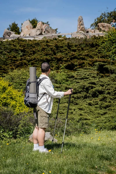 Вид на молодую коротковолосую женщину-туристку с рюкзаком и туристическим оборудованием, держащую в руках треккинговые шесты во время прогулки с пейзажем на заднем плане, любопытный турист, исследующий новые пейзажи — стоковое фото