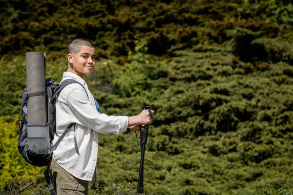 Sonriente joven excursionista de pelo corto con mochila sosteniendo bastones de trekking y mirando a la cámara con la colina y el paisaje en el fondo, Traducción del tatuaje: amor - foto de stock