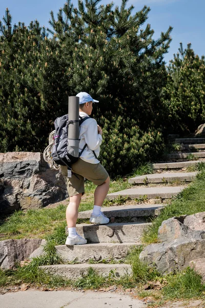 Молодая женщина-туристка в бейсболке и повседневной одежде с рюкзаком и туристическим оборудованием, идущая по лестнице с природой и небом на заднем плане, любопытный турист, исследующий новые пейзажи — стоковое фото