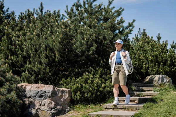 Jovem caminhante em roupas casuais e boné de beisebol segurando mochila enquanto desce escadas com a natureza e o céu azul no fundo, caminhante curioso explorando novas paisagens — Fotografia de Stock