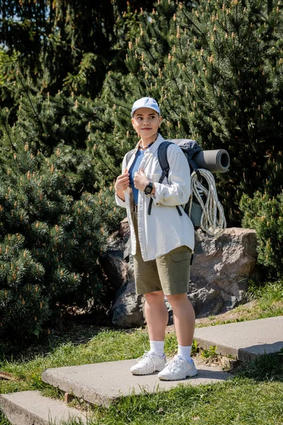 Позитивна молода жінка-мандрівник в бейсбольній шапці і повсякденному одязі тримає рюкзак і дивиться вбік, стоячи з природою на фоні, цікавий пішохід досліджує нові пейзажі — стокове фото