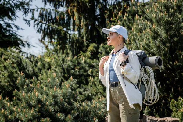 Side view of young short haired female tourist in casual clothes and baseball cap looking away while standing with blurred trees at background, curious hiker exploring new landscapes — Stock Photo