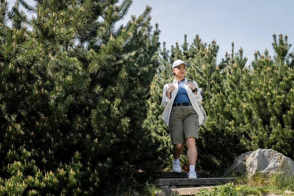 Young and short haired female hiker in casual clothes and baseball cap holding backpack and walking near trees with sky at background, woman trekking across vast landscapes — Stock Photo