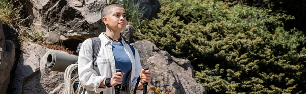 Young short haired female hiker in casual clothes with backpack holding trekking poles and looking away while standing with nature at background, woman trekking across vast landscapes, banner — Stock Photo