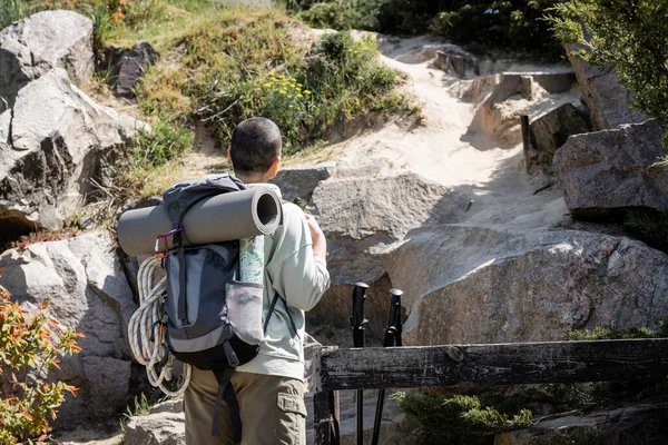 Rückenansicht einer jungen, kurzhaarigen Wanderin mit Rucksack, Fitnessmatte und Kletterseil im Hintergrund mit Hügel und Steinen, die durch weite Landschaften wandert — Stockfoto