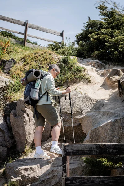 Vista lateral de la joven viajera femenina de pelo corto en ropa casual con mochila sosteniendo bastones de trekking mientras camina sobre una colina con piedras durante el verano, Traducción del tatuaje: amor - foto de stock