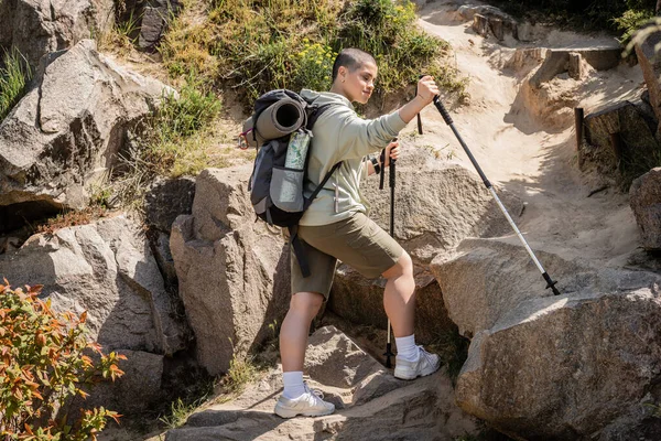 Young short haired female traveler in casual clothes with backpack holding trekking poles while standing near hill with stones at background during summer, Translation of tattoo: love — Stock Photo