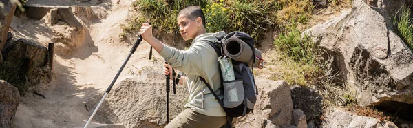 Short haired woman traveler in casual clothes with backpack holding trekking poles while standing near hill with stones at background, tranquil hiker finding inner peace on trail, banner — Stock Photo